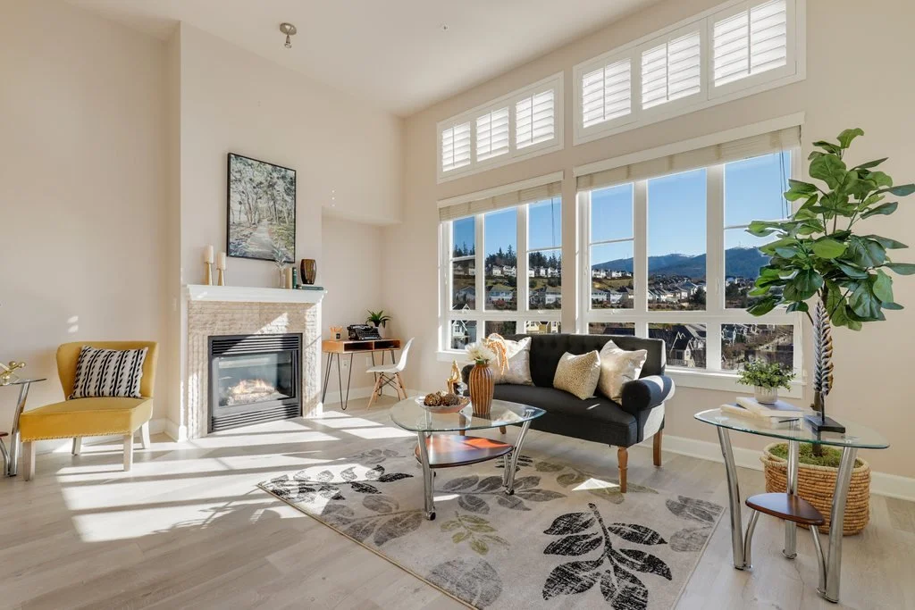 Bright living room with large windows showing a mountain view, a black sofa with beige pillows, a glass coffee table with decor, a yellow chair, a potted plant, and a fireplace with artwork above it.