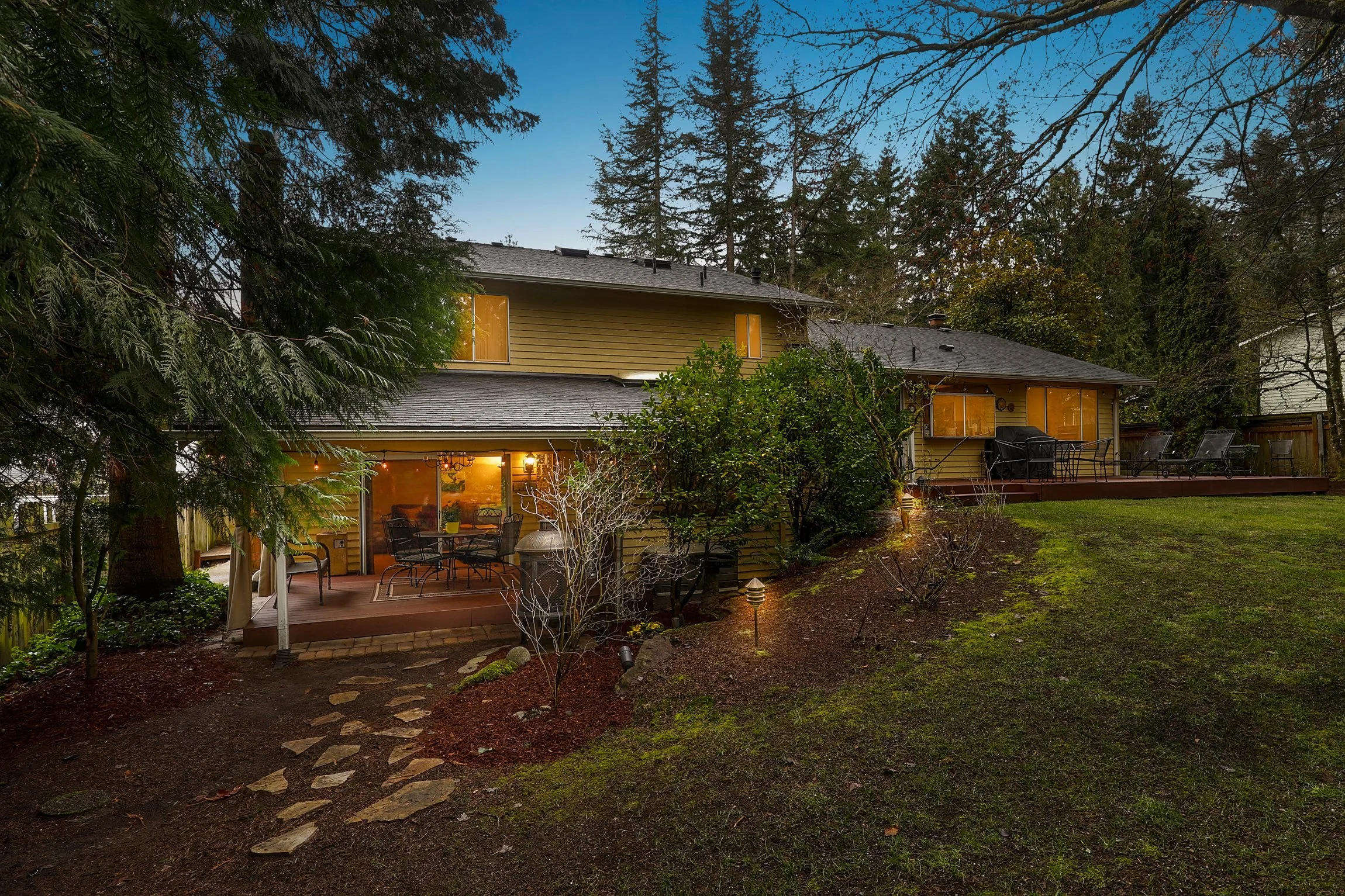 Backyard view of a yellow house with a deck and outdoor furniture, surrounded by tall trees and a well-maintained lawn during dusk.