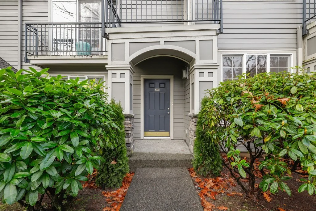 Front entryway of a residential building with a gray door, surrounded by green bushes and small trees, with a walkway leading up to it.