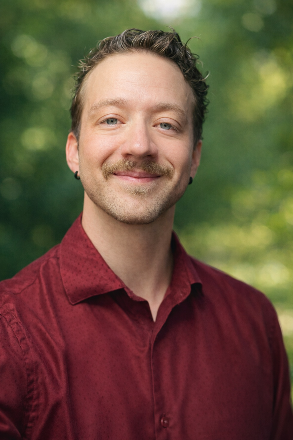 A smiling man with light skin, mustache and beard, short curly hair, wearing black earrings and a red shirt, standing outdoors with a blurred green background.