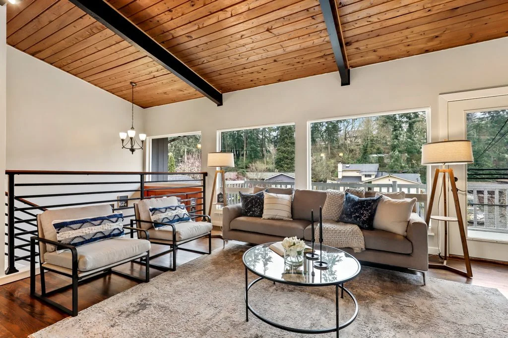 Living room with wooden vaulted ceiling, large windows, beige sofa, two armchairs with black frames, a round glass coffee table, and floor lamps, overlooking a neighborhood with trees and houses.
