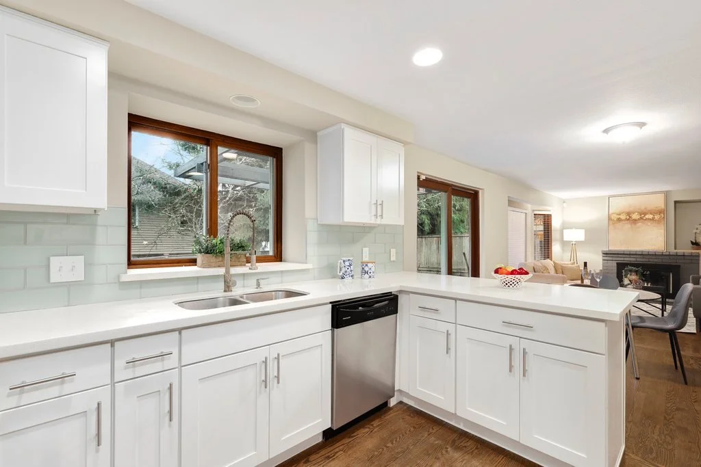 Modern kitchen with white cabinets, green subway tile backsplash, stainless steel dishwasher, and a large window above the sink. Open living space visible in the background.