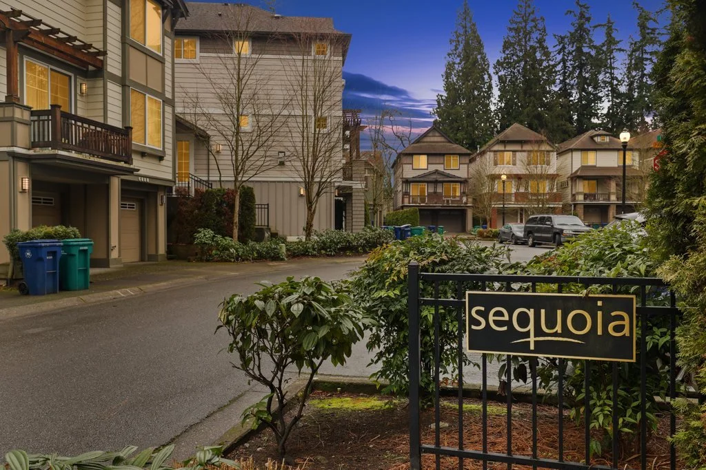 A residential street at dusk with townhouses, trees, and parked cars. A sign reads "Sequoia," and trash bins are visible near the curb.