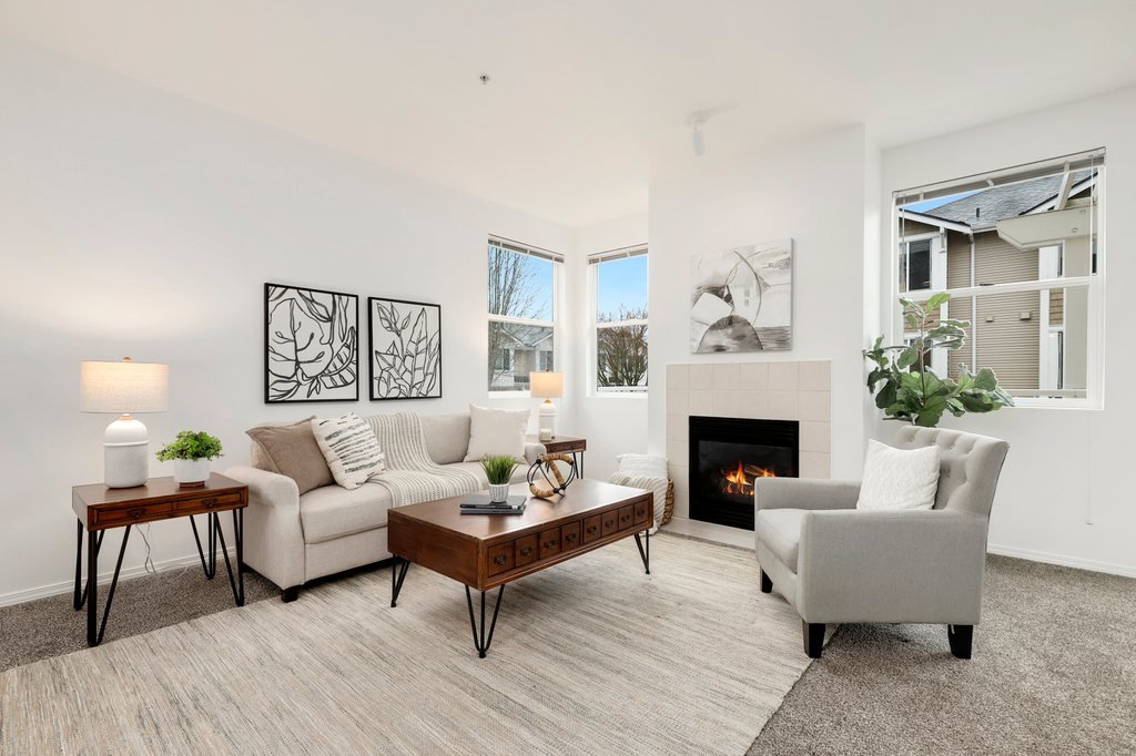 A modern living room featuring a white sofa with throw pillows, a white armchair, a wooden coffee table, two side tables with lamps, a fireplace, and artwork on the walls, with windows showing neighboring houses.