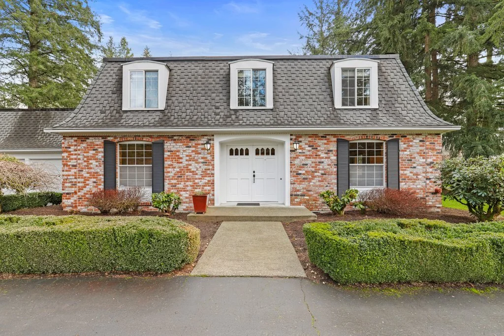 Front view of a brick house with dormer windows and a white door, surrounded by manicured bushes and trees under a cloudy sky.