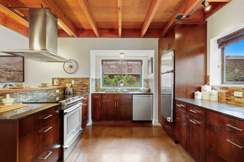 A modern kitchen with wooden cabinets and a wooden ceiling, stainless steel appliances, tiled backsplash, and two windows providing natural light.