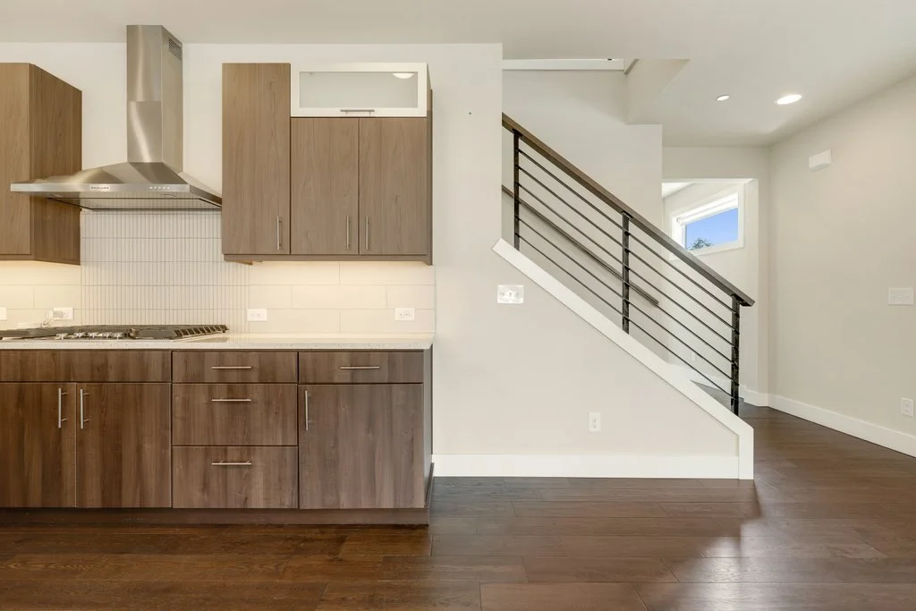 Modern kitchen with wooden cabinets, stainless steel range hood, white tile backsplash, and a staircase with black rails leading upstairs, hardwood floors, and white walls.