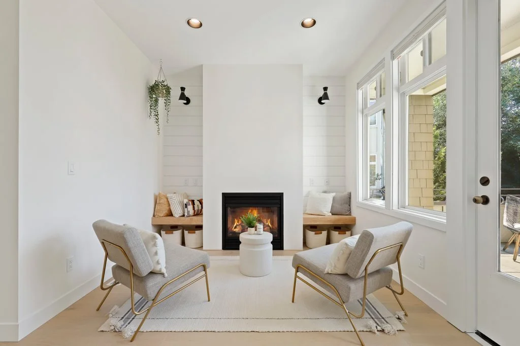 Bright living room with a centered fireplace, two armchairs facing each other, large windows on the right, small round table with a plant in front, and window seats with cushions on either side of the fireplace.