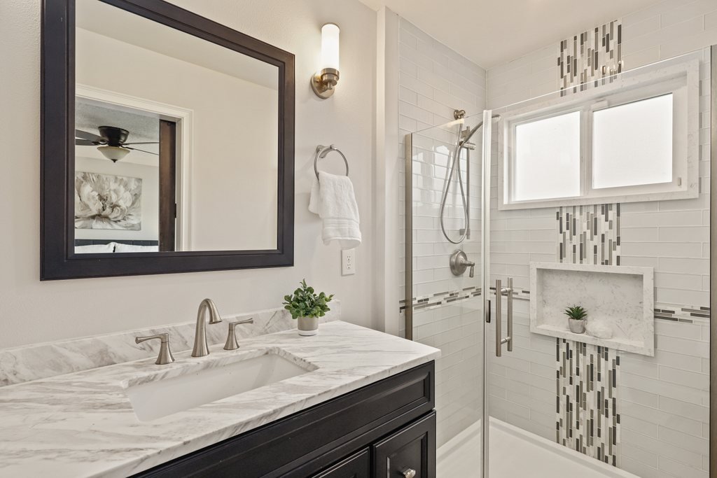 Modern bathroom with marble countertop sink, dark wood vanity, large mirror, glass shower enclosure with built-in shelf and window, white tile walls with decorative vertical tile strip, small potted plant on countertop, towel hanging on wall, and a w