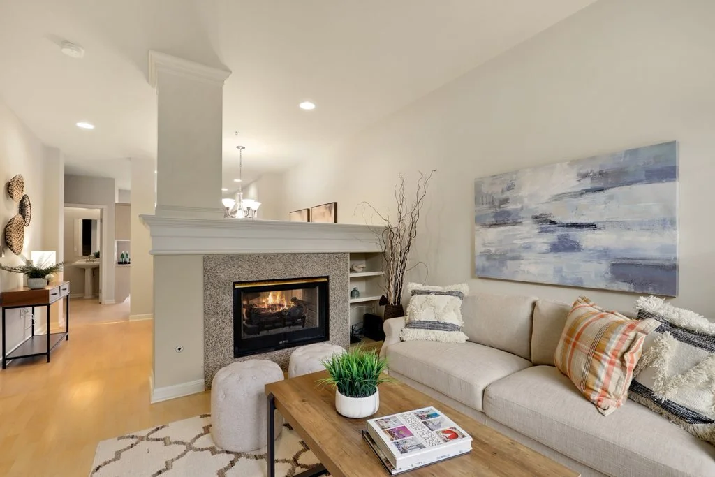 Living room with beige sectional sofa, patterned pillows, abstract wall art, fireplace, wooden coffee table with magazine and plant, and decorative wall accents.