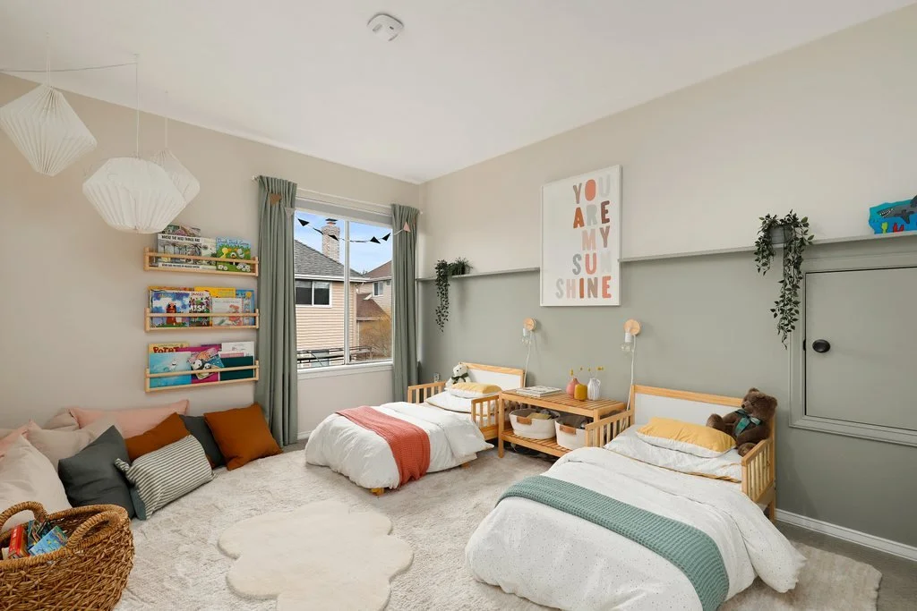 A bright children's bedroom with two beds, a window, bookshelves, and plush pillows. Light-colored walls with hanging paper lanterns and plant decorations.