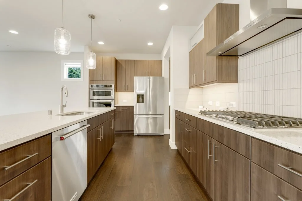 Modern kitchen with wooden cabinets, white countertops, stainless steel refrigerator, double oven, and gas stove, with pendant lighting and small window.