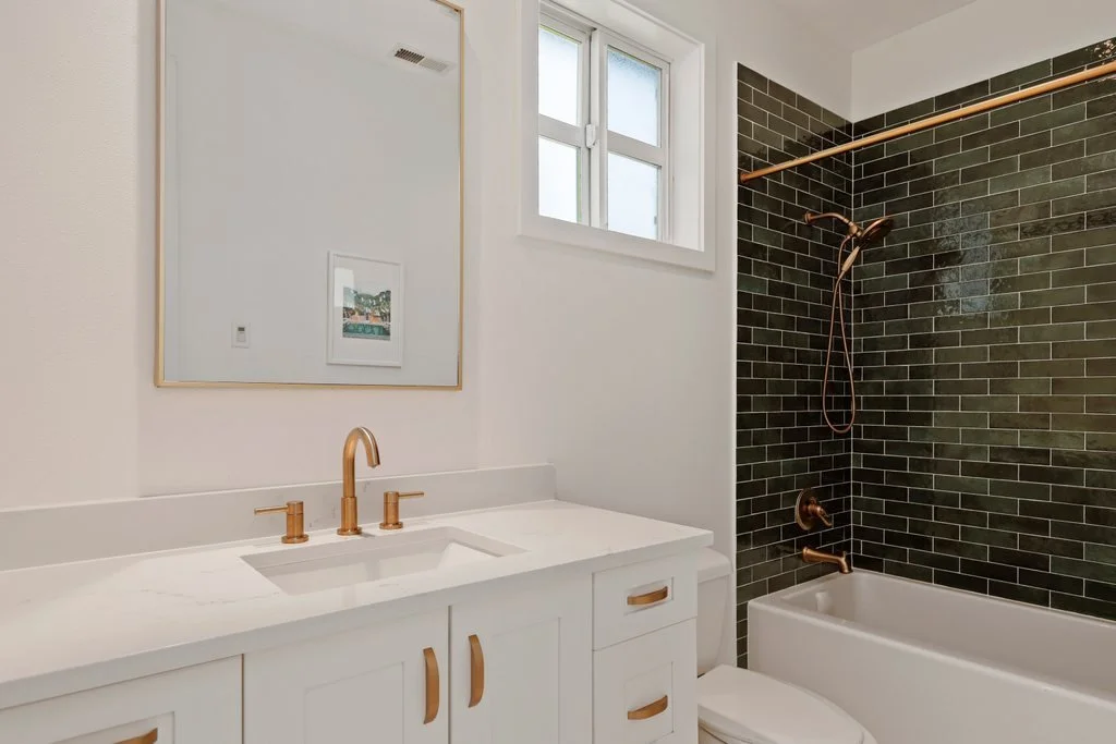 Modern bathroom with white vanity, gold fixtures, large mirror, and dark tile shower wall with bronze fixtures