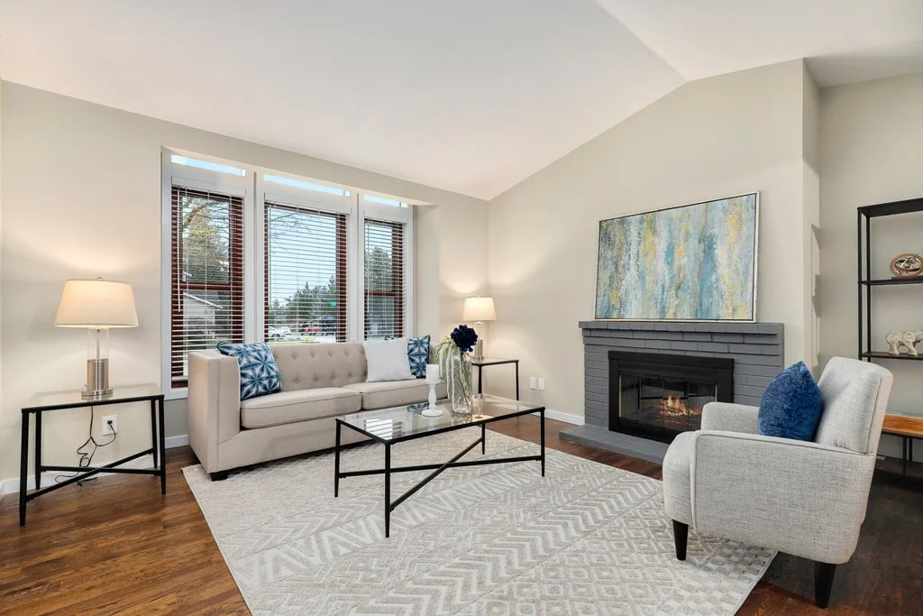 Living room with beige sofa, white armchair, fireplace, three windows with blinds, side tables with lamps, and decorative pillows.