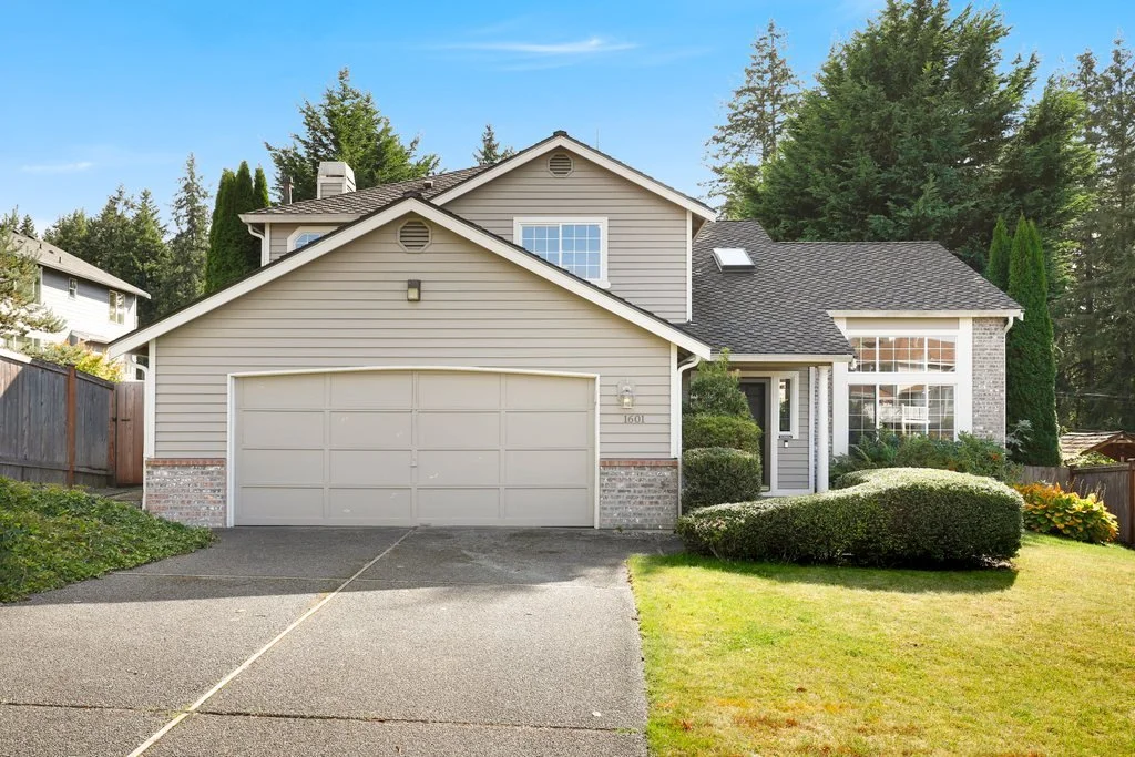 Front view of a two-story house with a garage, surrounded by a well-maintained lawn and trees under a clear sky.