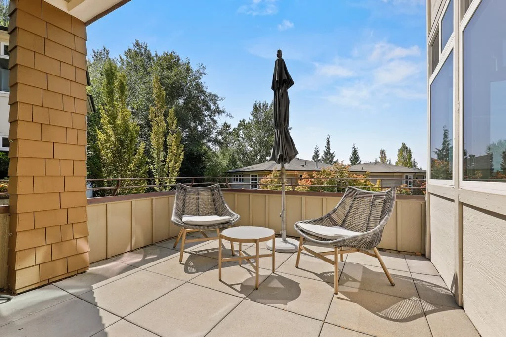 Empty balcony with two modern chairs, a small table, and a closed umbrella, overlooking trees and neighboring houses under a clear blue sky.