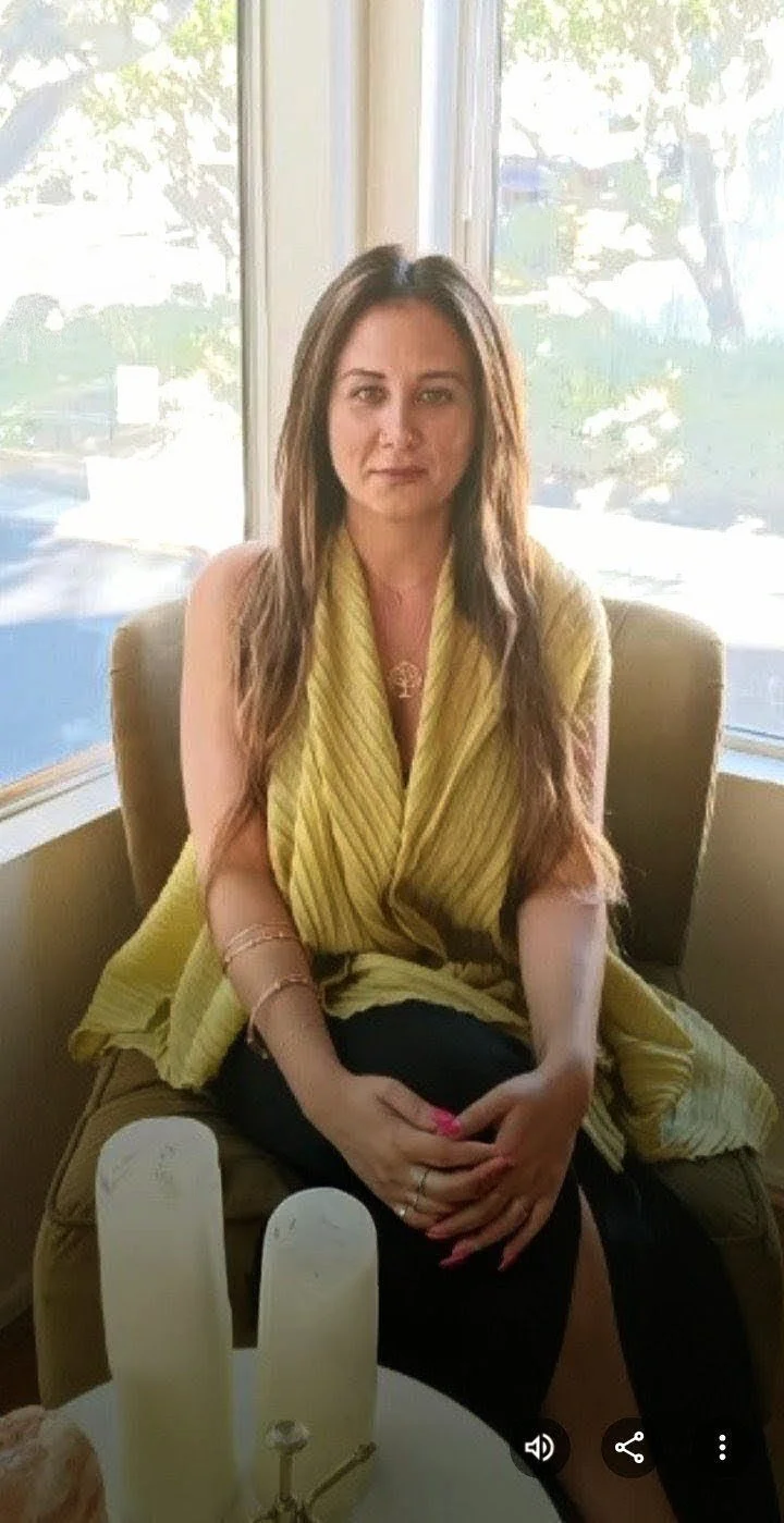 A woman with long brown hair sitting on a beige armchair by a window, wearing a yellow sleeveless top and black pants, with her hands clasped on her lap.