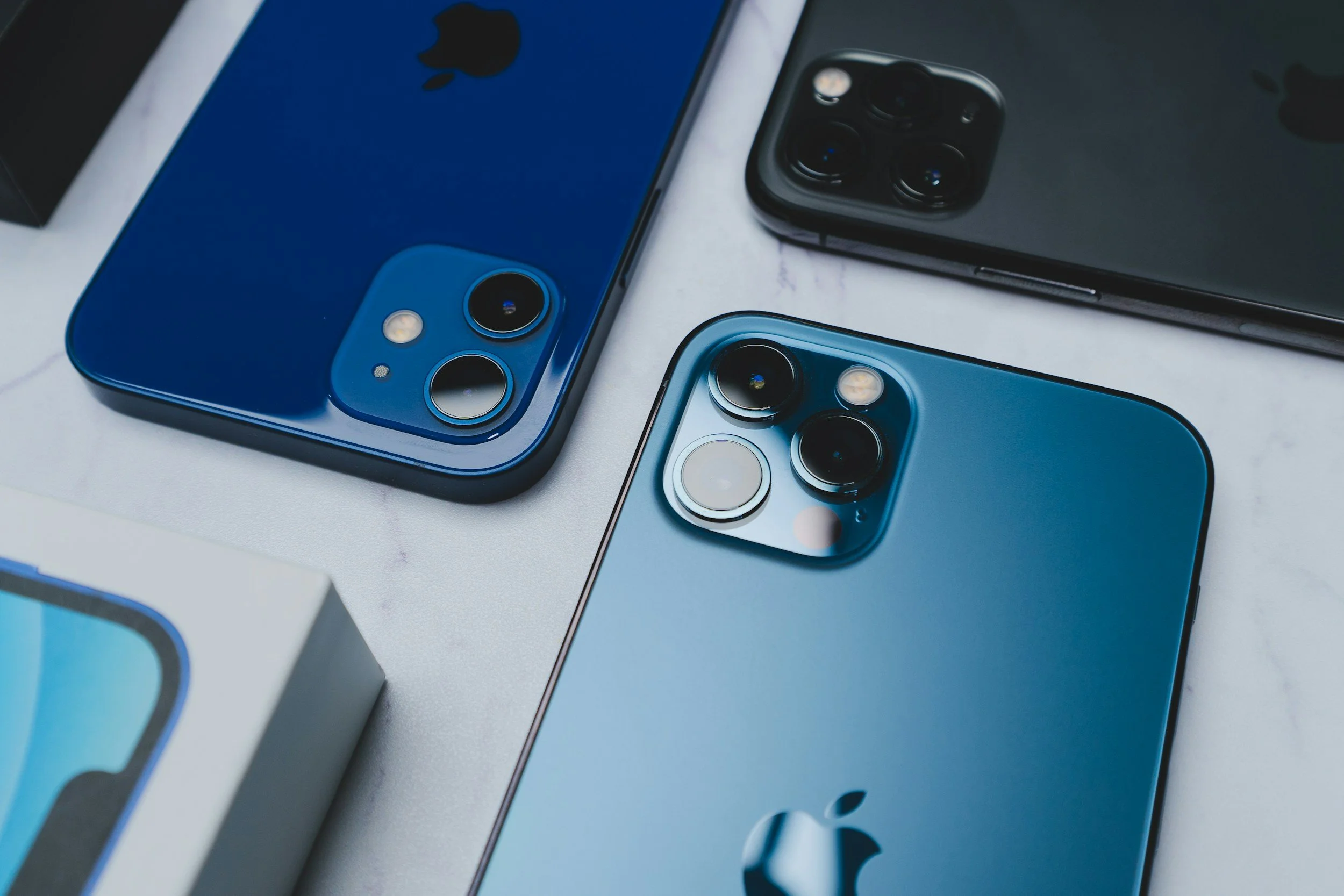 Close-up of three iPhones with triple and dual camera setups, in blue, black, and teal colors, on a white surface.
