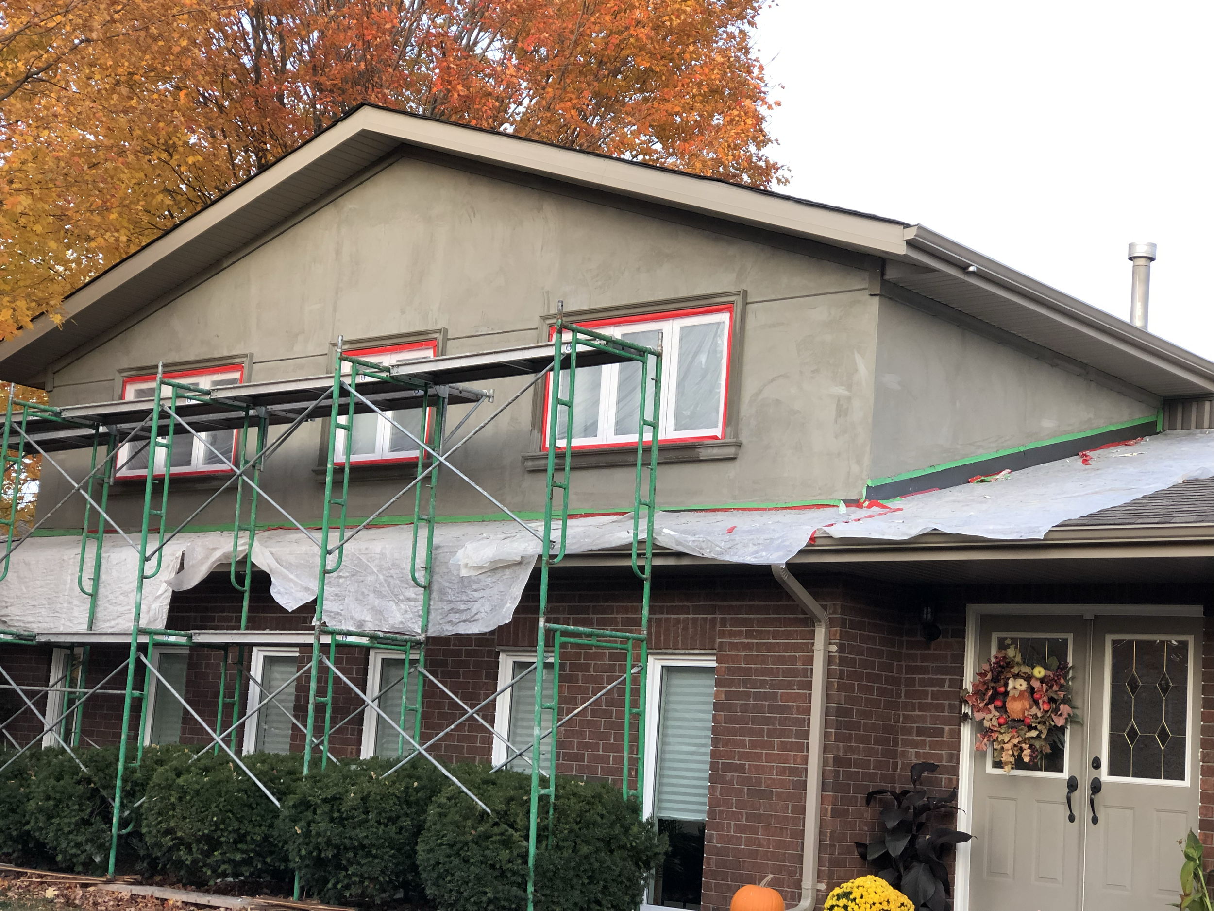 Scaffolding set up against the side of a house under an orange autumn tree, with some window trim taped red.