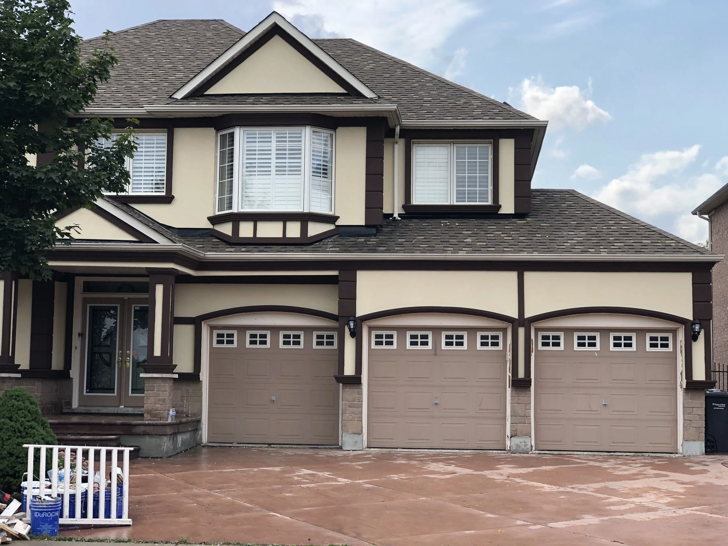 Front view of a two-story house with a beige and brown color scheme, featuring a three-car garage with decorative windows, a main entry door, and a large bay window on the second floor. There is a tree on the left side and some painting supplies on t