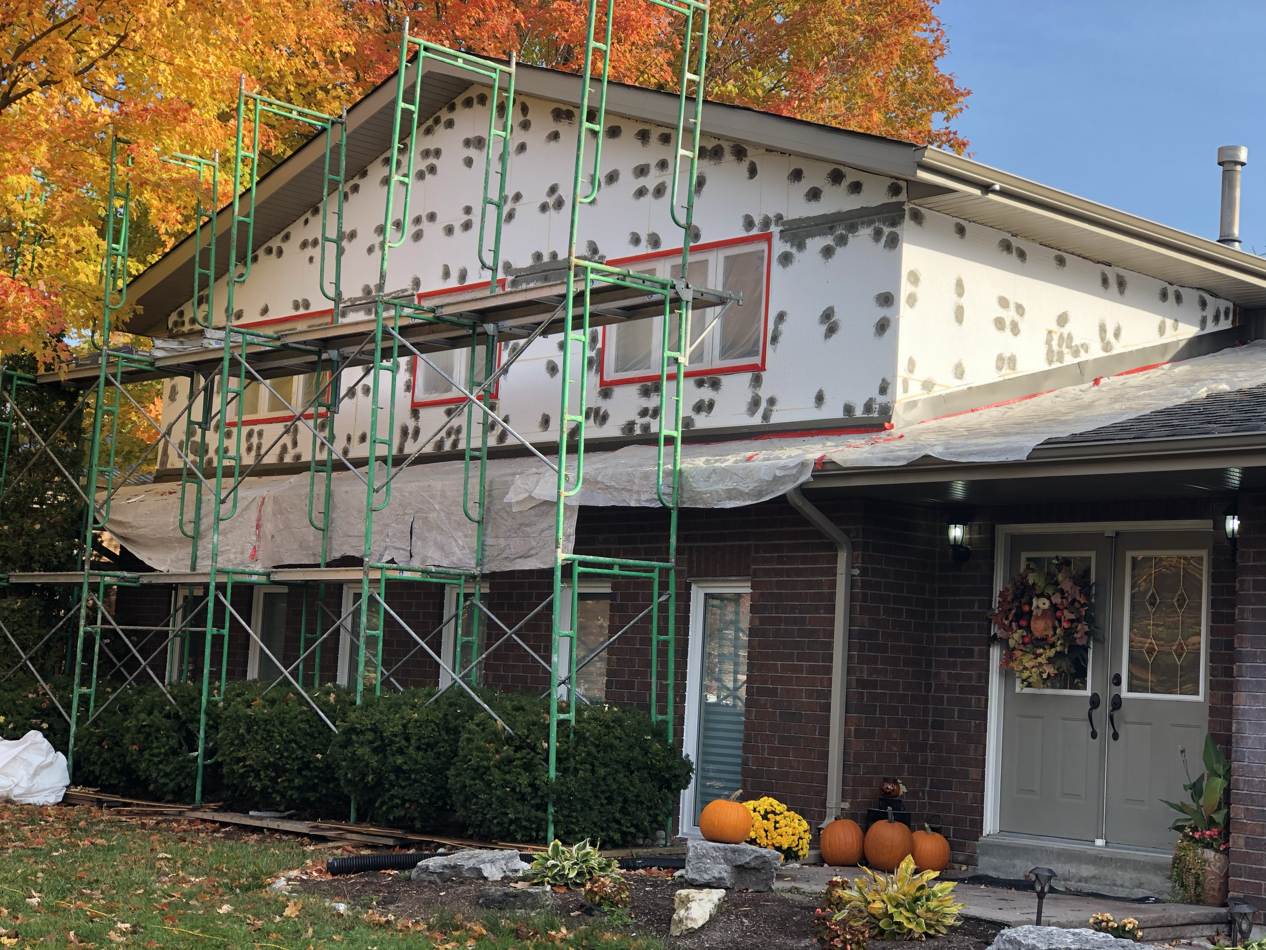 House under renovation with scaffolding, black spots on white exterior walls, fall foliage in the background, pumpkins and yellow flowers outside, and a large window with red trim.