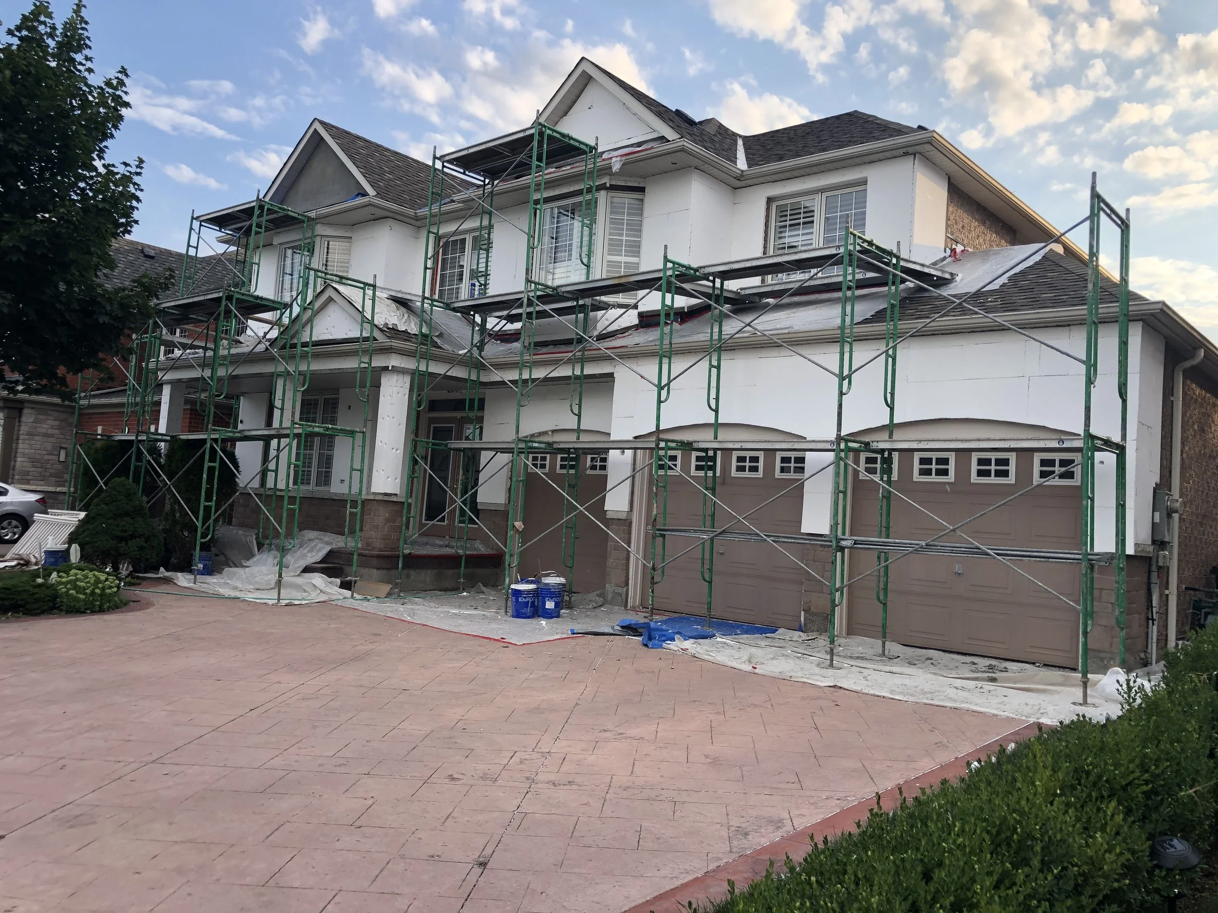 A house undergoing roof renovation with green scaffolding, white walls, a gray garage door, and a paved driveway, with construction materials and tools around.