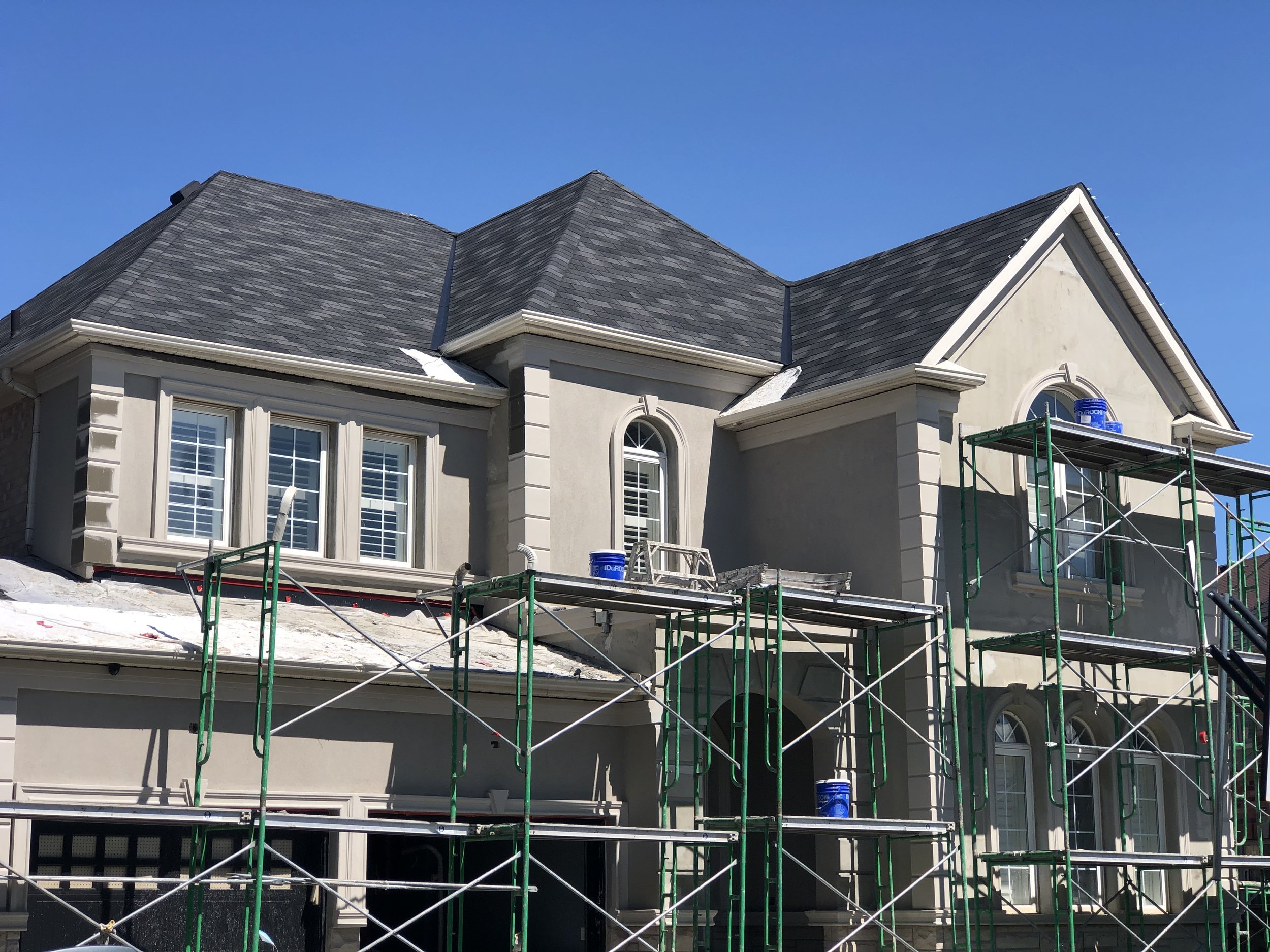 A house with beige stucco walls and white trim, under construction or renovation, with scaffolding surrounding it and a clear blue sky overhead.