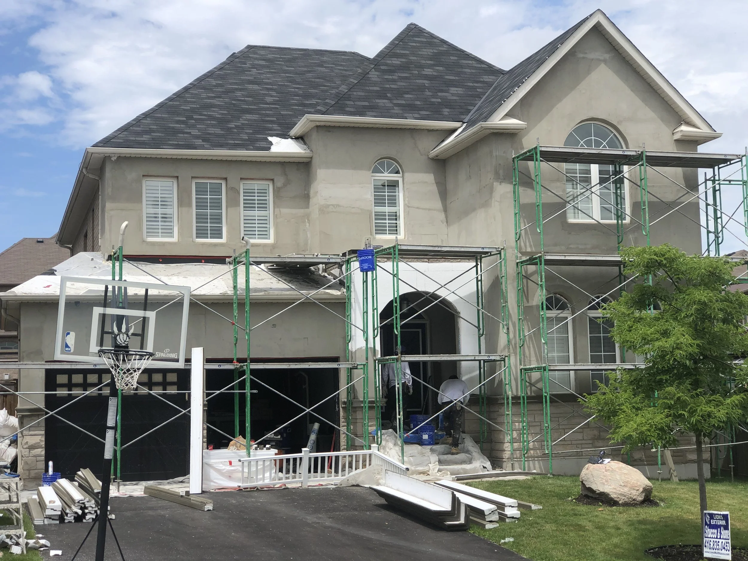 A house under construction with scaffolding along the front and workers working on the exterior. The house has a new roof and unfinished walls. There's a basketball hoop on the driveway, and construction materials and equipment scattered around.