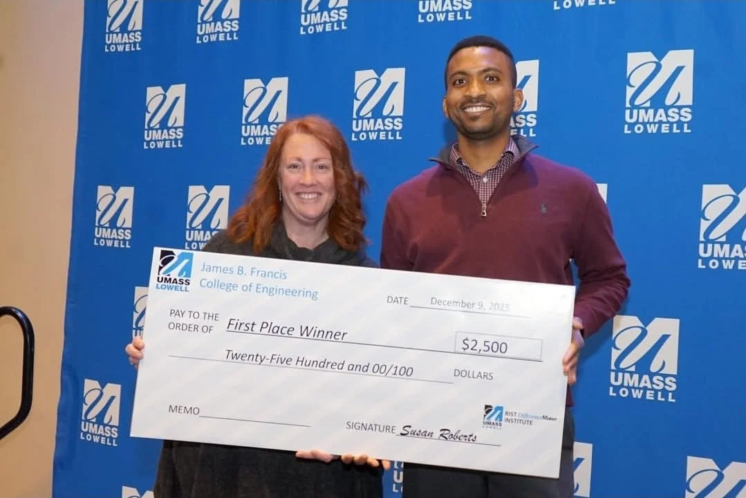 Two individuals, a woman with red hair and a man with dark hair, smiling and holding a large check for $2,500 at an event with a blue UMASS Lowell backdrop.