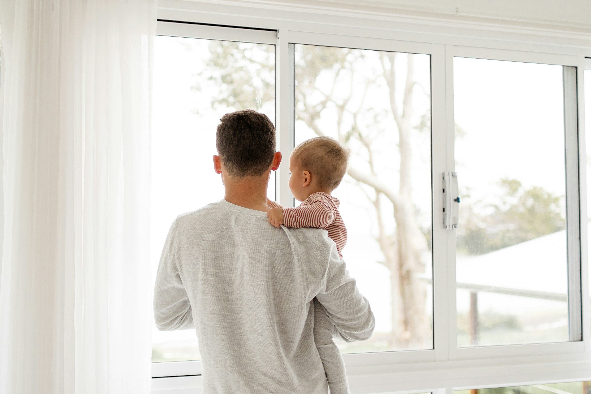 An adult holding a young child while looking out a large window with a white frame, revealing trees and sky outside.