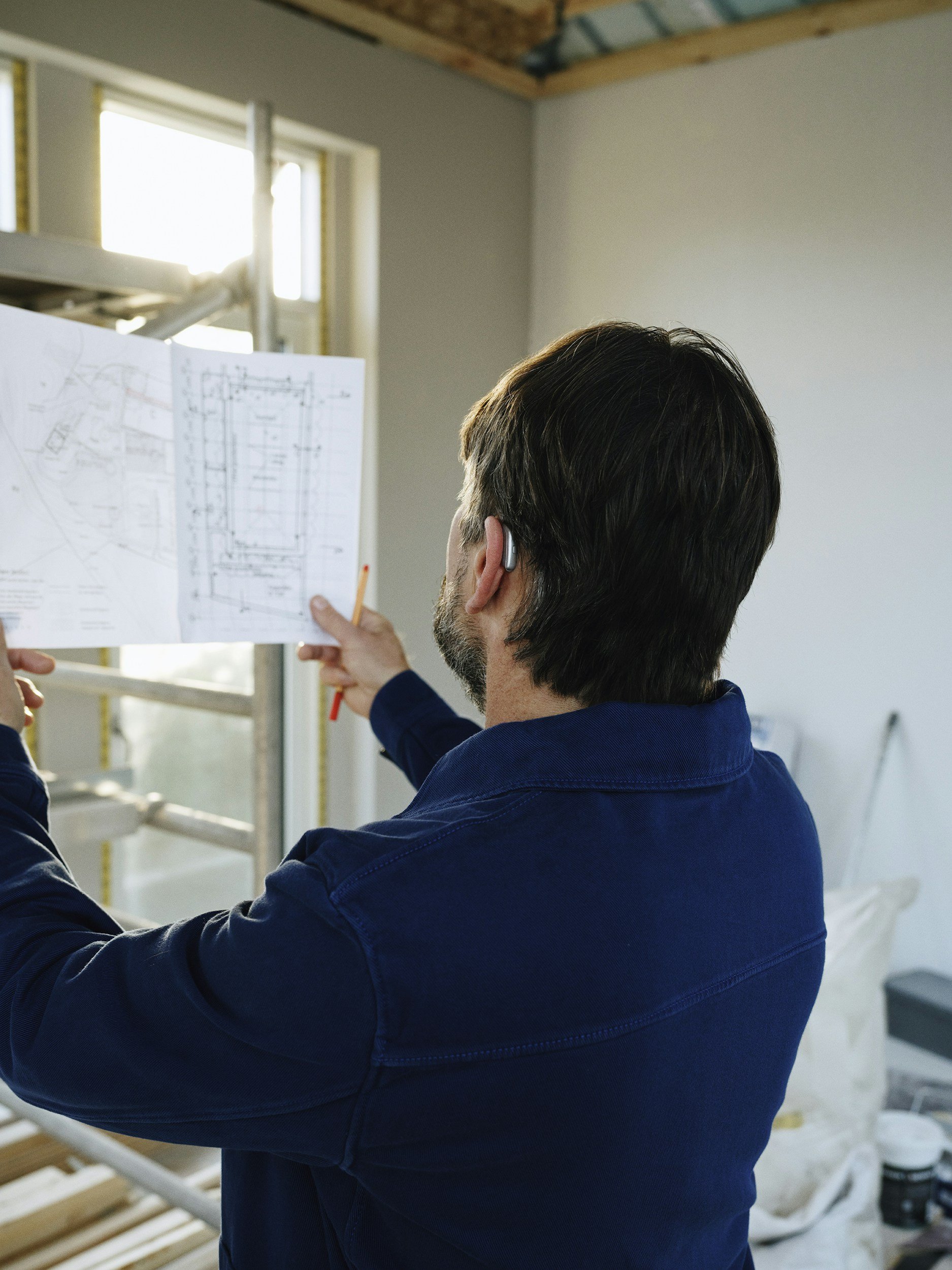A man with dark hair and beard, wearing a blue shirt with rolled-up sleeves, is holding a sheet of paper with architectural drawings on it and a pencil, standing in a room under construction with visible framing and a window.