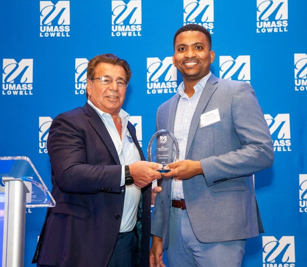 Two men in suits standing in front of a blue UMASS LOWELL backdrop, smiling as one presents an award to the other during a ceremony.