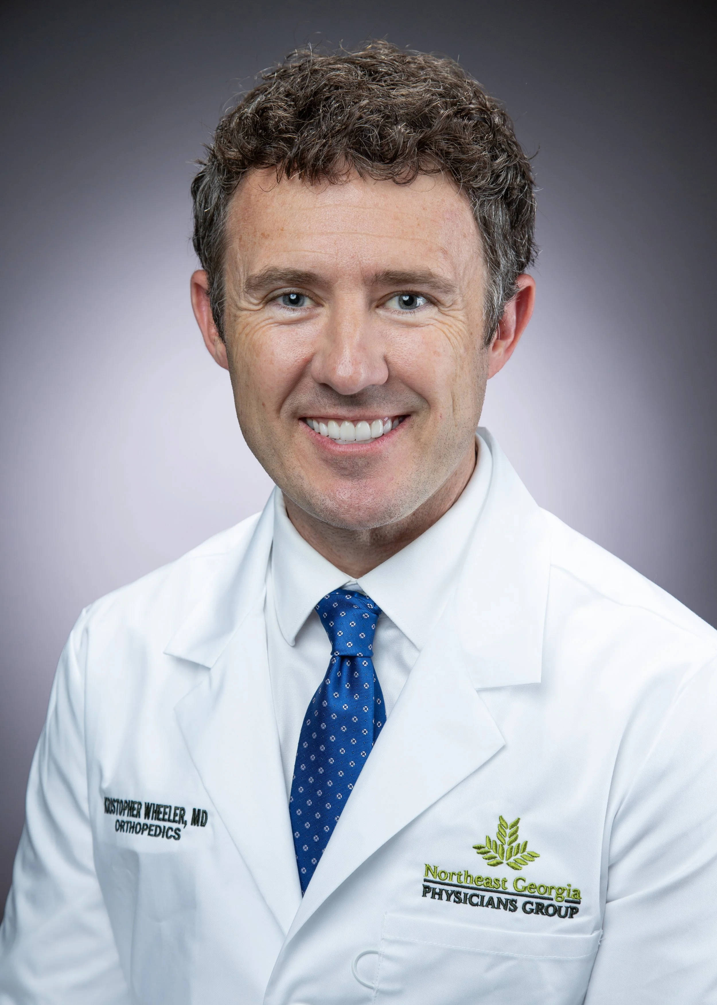 A smiling man in a white medical coat with hospital logo, dressed in a blue tie, against a neutral background.