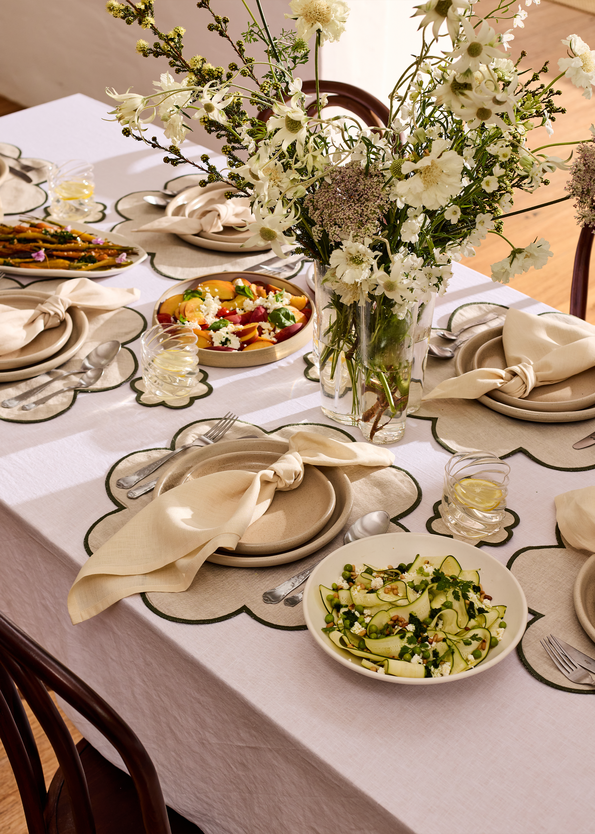 A dining table set with beige plates, silverware, beige cloth napkins, and circular placemats with black edges. There is a large bouquet of white and light purple flowers in a glass vase at the center. The table features bowls of salad, including one with zucchini, peas, and herbs, and another with fruit and cheese. Small glass bowls of lemon water are placed near each setting.