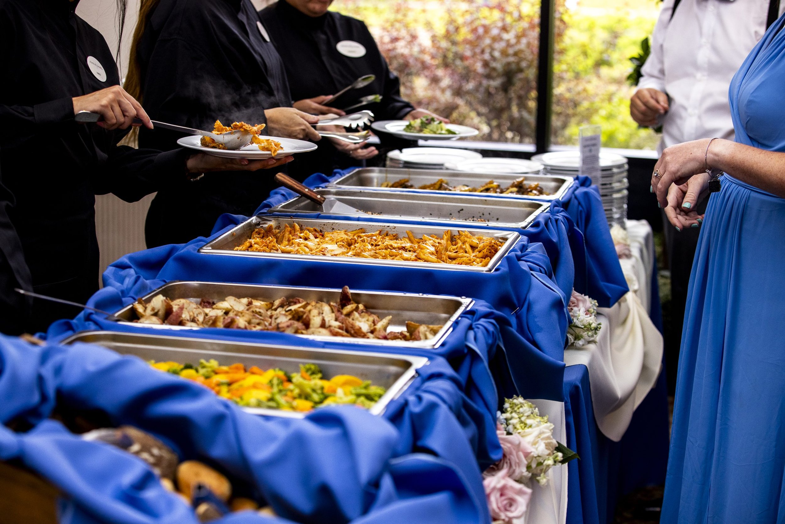 Buffet table with various dishes including pasta, roasted vegetables, and meat, covered with blue and white tablecloths, at a social gathering.
