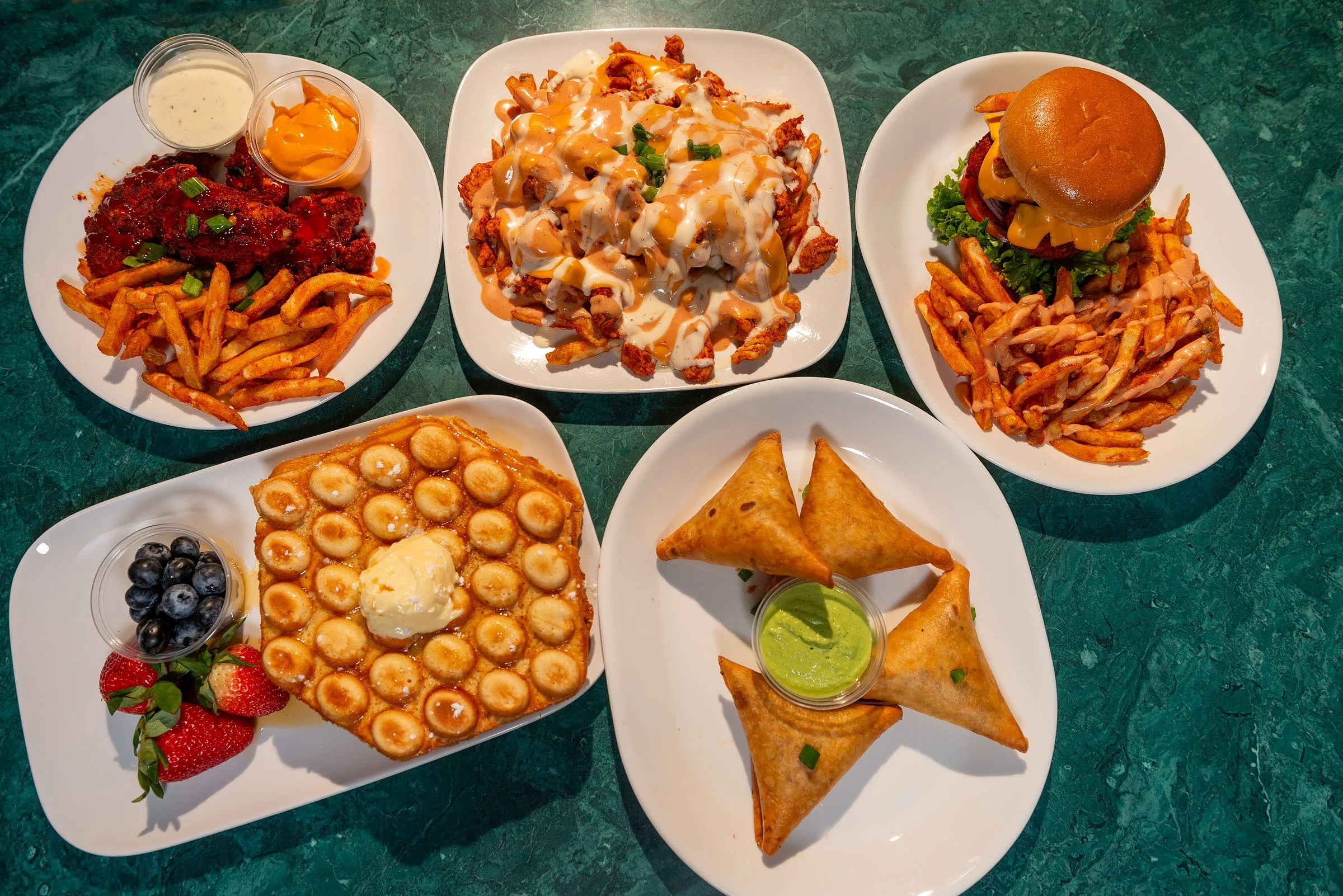 A variety of American diner food dishes on a green marble table, including chicken wings with sauce, a plate of poutine with gravy and cheese curds, a hamburger with fries, a dessert topped with mini cream pies and ice cream, a plate of samosas with green sauce, and a plate of fries with ranch and buffalo sauces.