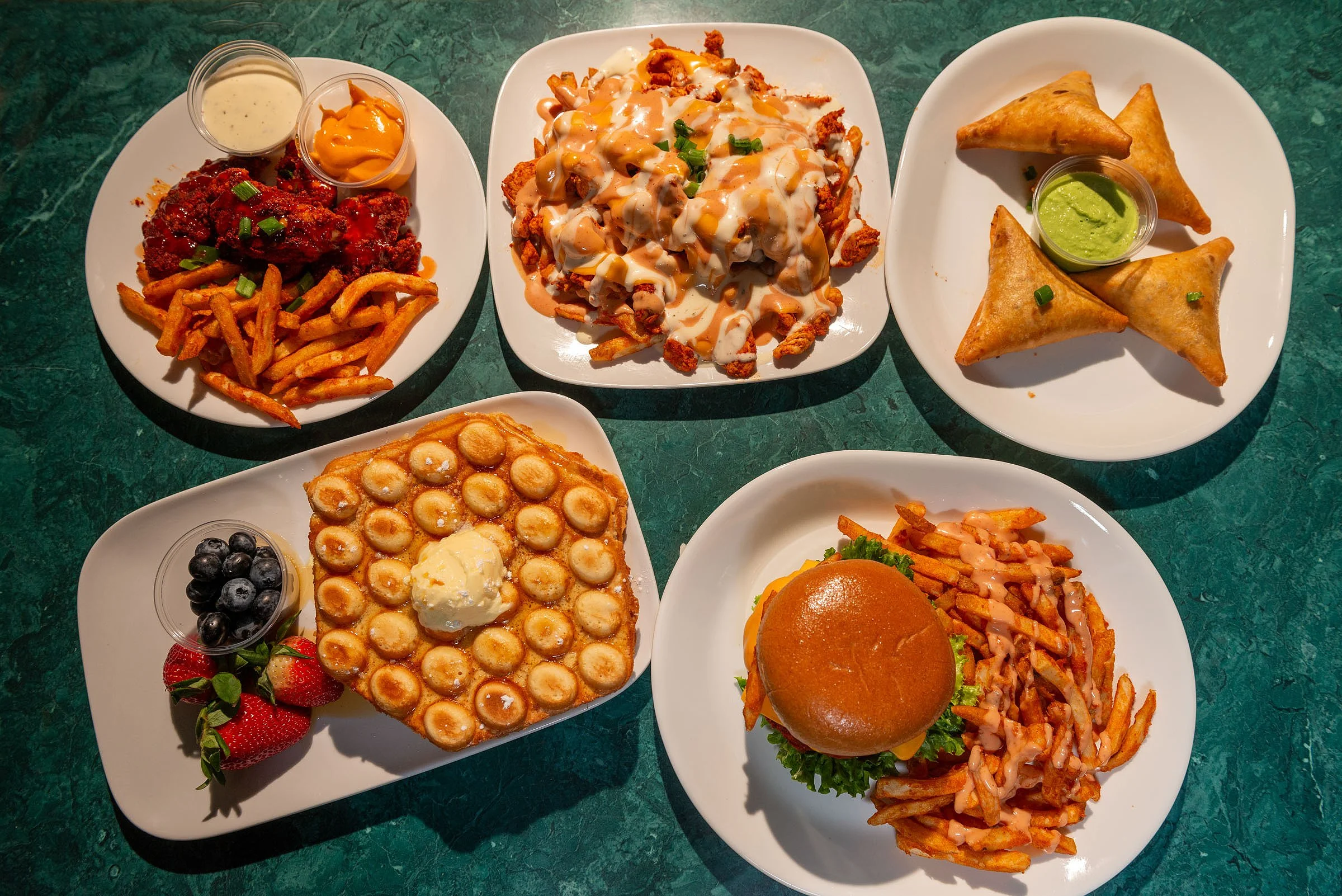 Assorted plates of American comfort food including chicken wings with two sauces and fries, loaded nachos with cheese and gravy, fried wontons with dipping sauce, mini chicken pot pie with berries, and a burger with fries and sauce.
