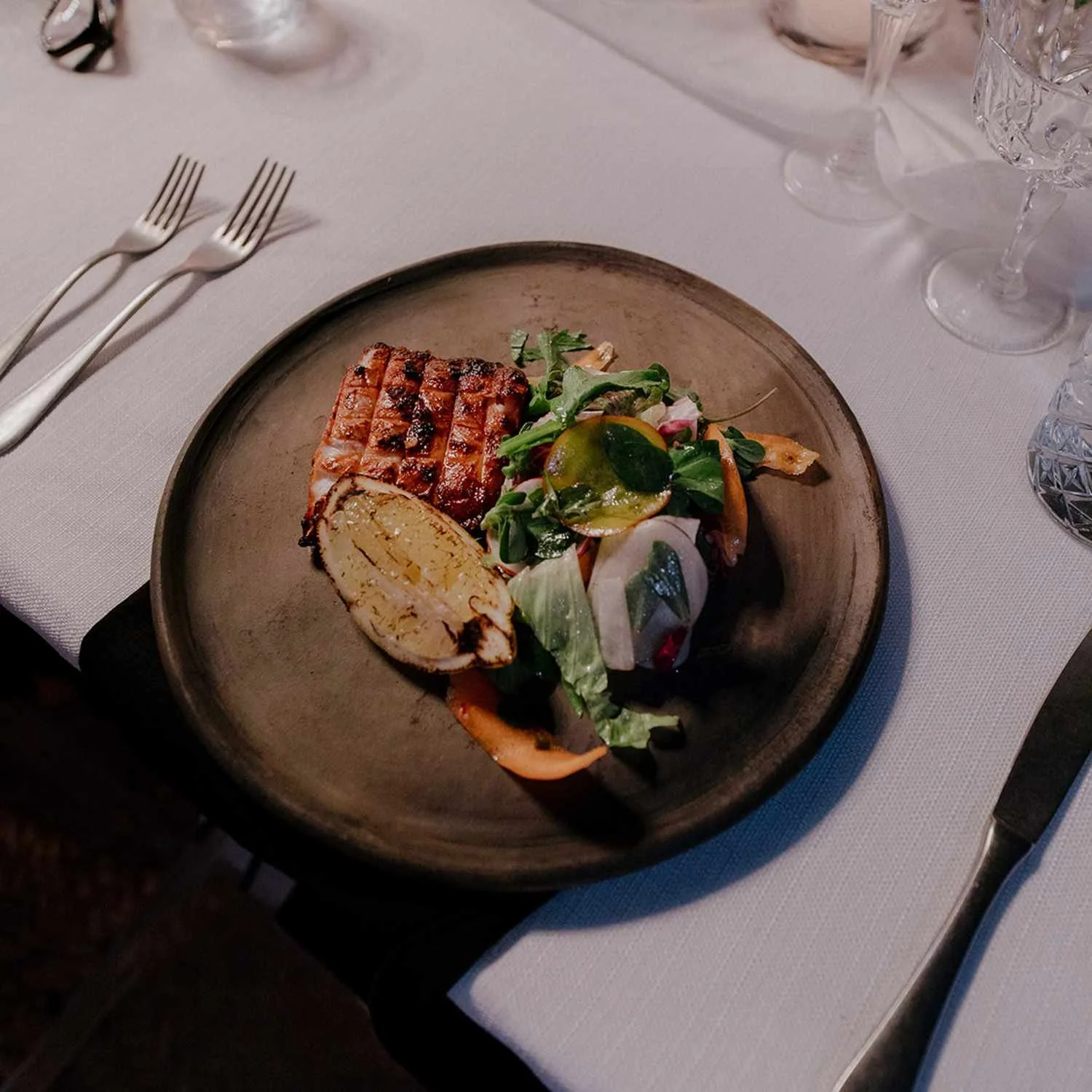 A plate of grilled meat, a stuffed chicken breast, and a small salad with greens and dressing on a dark ceramic plate, set on a white tablecloth with silverware and glassware around.