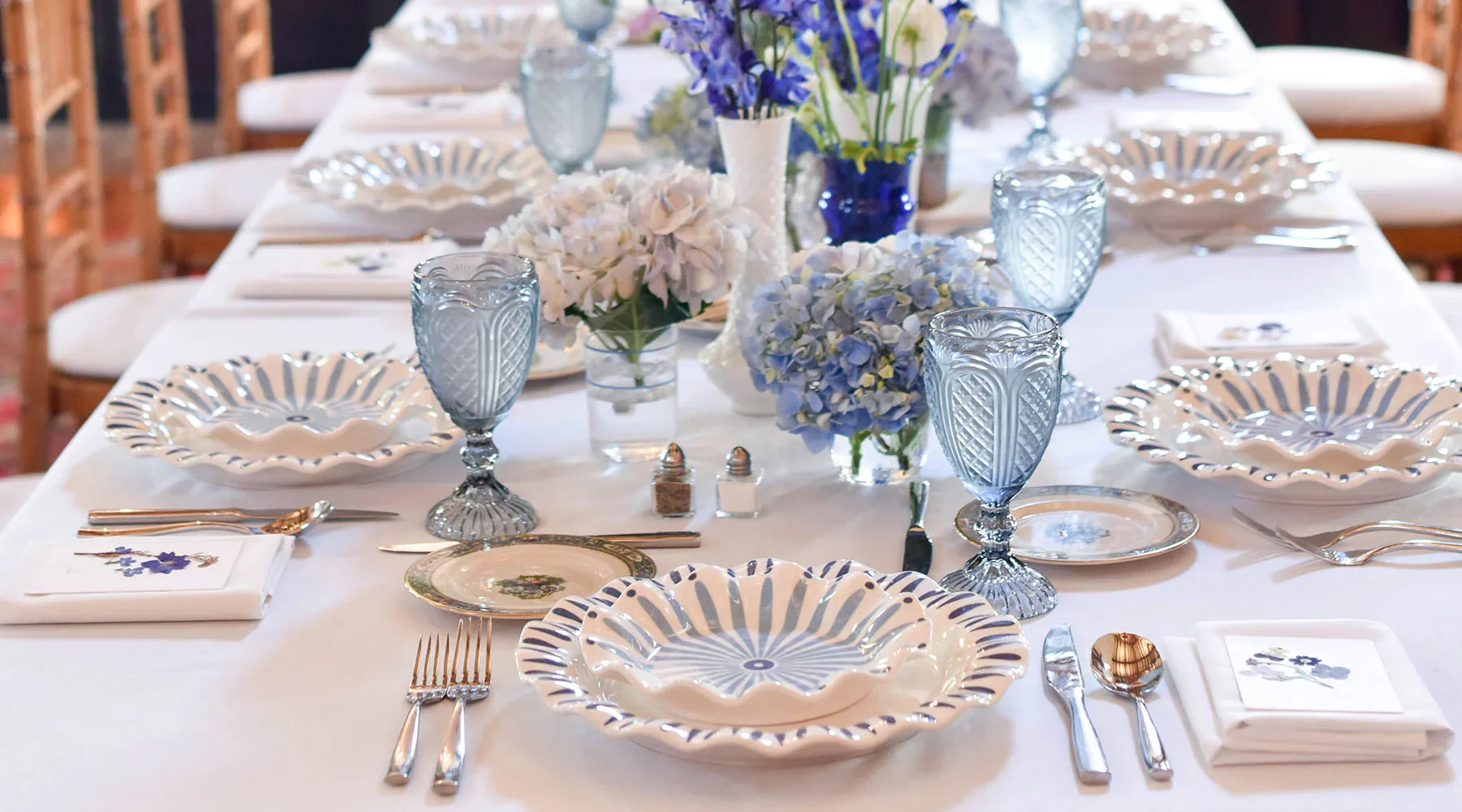 Elegant dining table set with blue and white patterned porcelain plates, matching blue glass goblets, silver cutlery, white napkins with floral embroidery, and floral centerpieces with white and blue hydrangeas and other flowers.