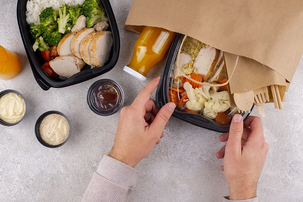 A person's hands opening a black takeout container filled with a salad containing chicken, broccoli, and other vegetables, on a light gray surface. Surrounding are small cups of various sauces, a bottle of mustard, a jar of barbecue sauce, and a bottle of orange juice.