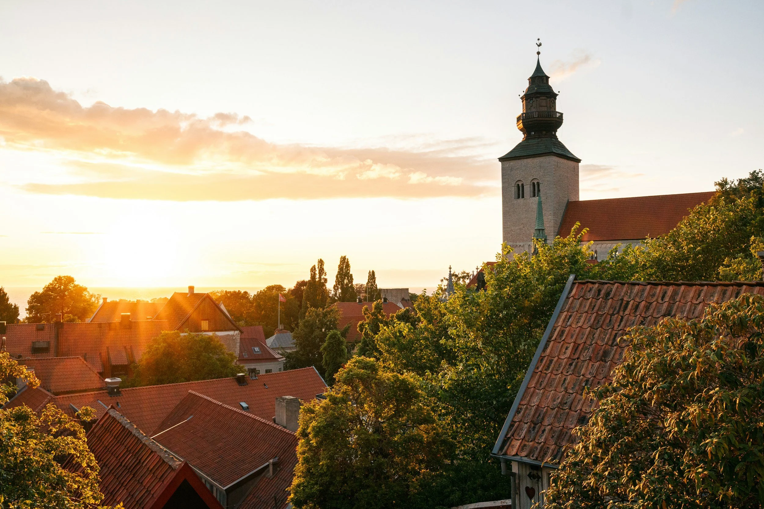 Ett foto på hustak och en kyrka i Visby, med kvällssolen nedåtgående i bakgrunden.