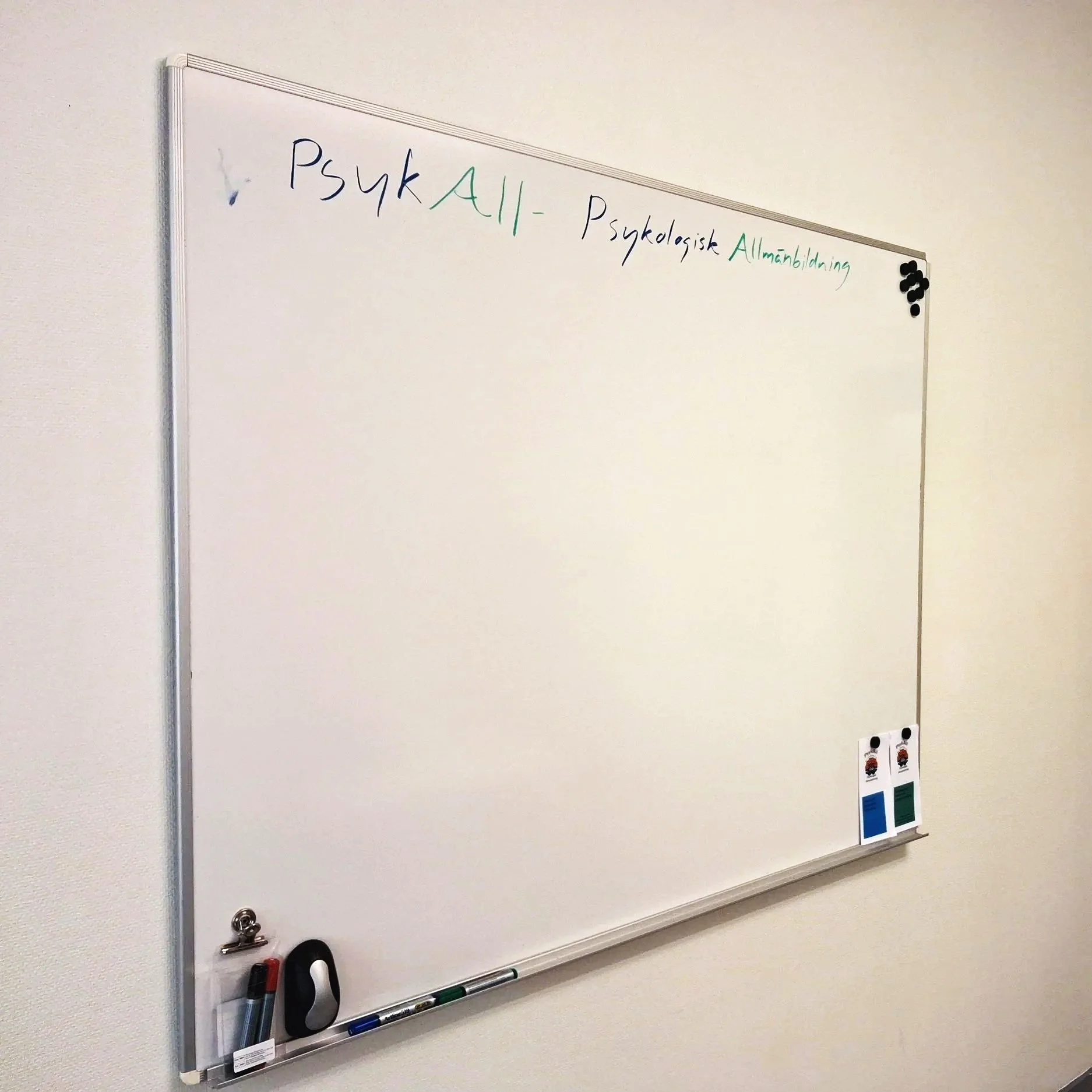 Foto av en whiteboard. Överst på tavlan står skrivet "PsykAll - Psykologisk Allmänbildning" i blå och grön penna. På nedre delen av tavlan finns pennor, en whiteboard-sudd, och två informationsbroschyrer om PsykAll
