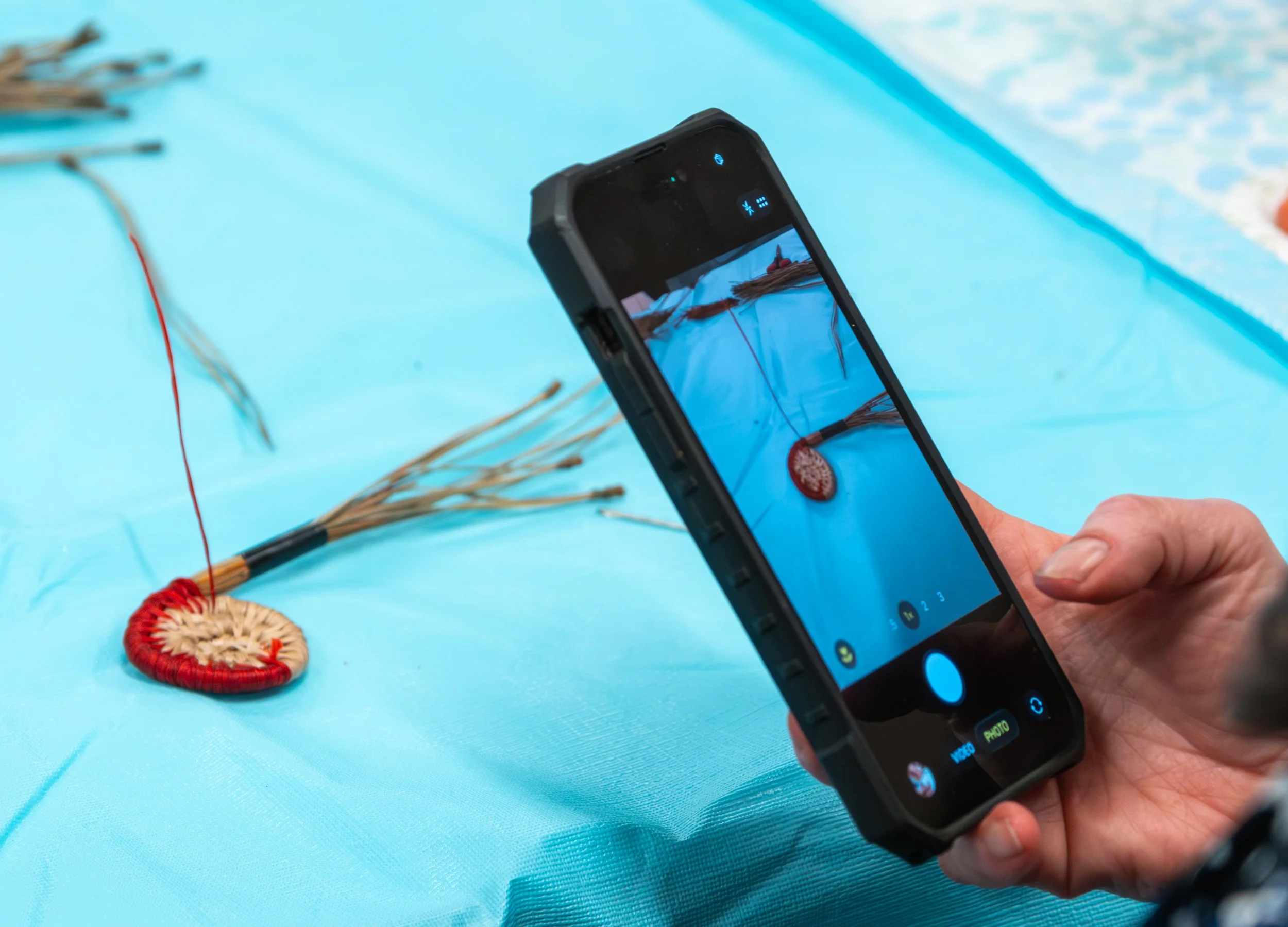 Close-up of a hand holding a smartphone taking a photo of a circular pine needle coaster weaving with red thread. The weaving is on a light blue table cloth.