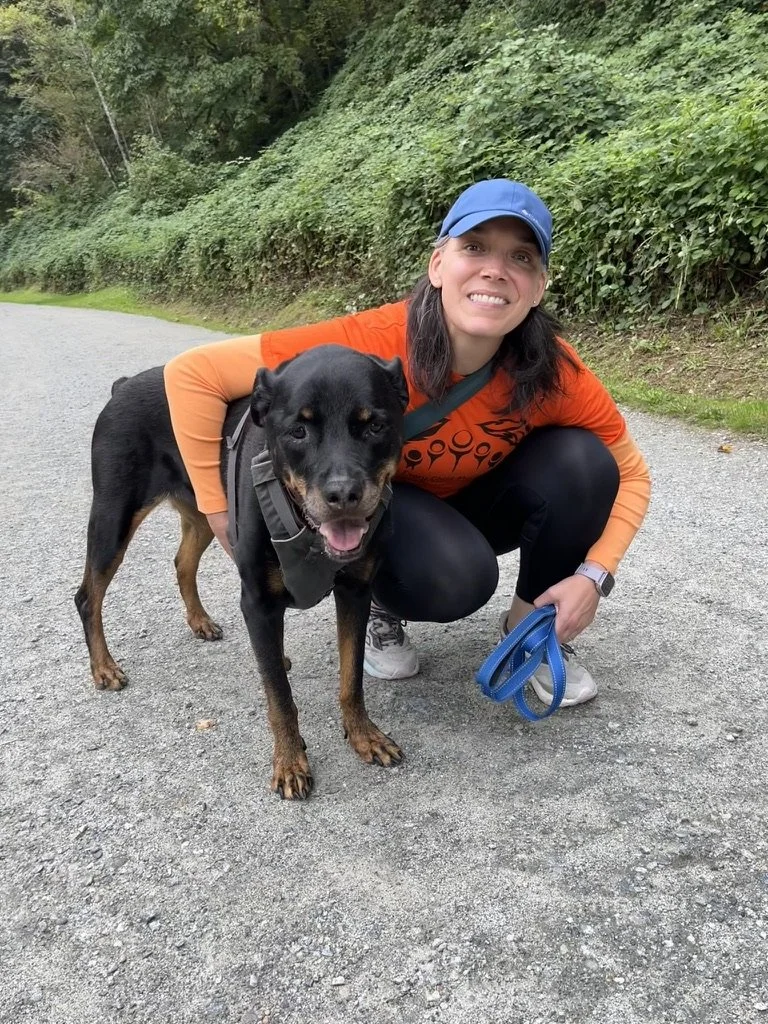 A woman with dark brown hair, wearing an orange Every Child Matters shirt, black pants, and a blue cap, crouches beside a black and tan dog on a gravel trail in a wooded area, smiling at the camera.