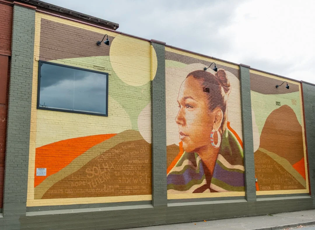 Mural on a brick building featuring a profile of a woman with a bun hairstyle, large hoop earrings, and colorful clothing, with abstract landscape background.