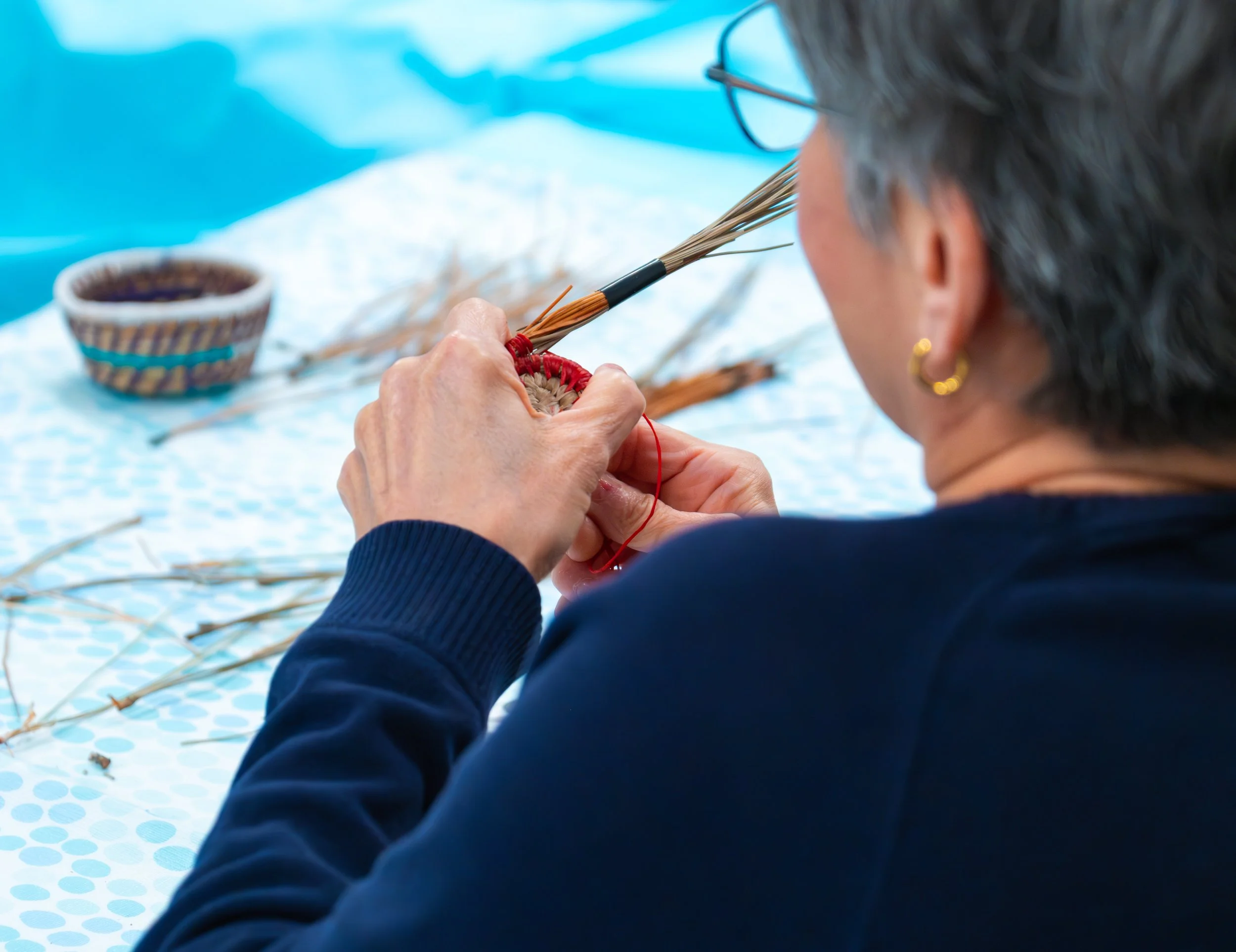 A person with short gray hair and wearing a dark blue sweater is weaving a pine needle coaster using red thread. In the background, there is a small woven basket and extra pine needles on a blue polka dot tablecloth.