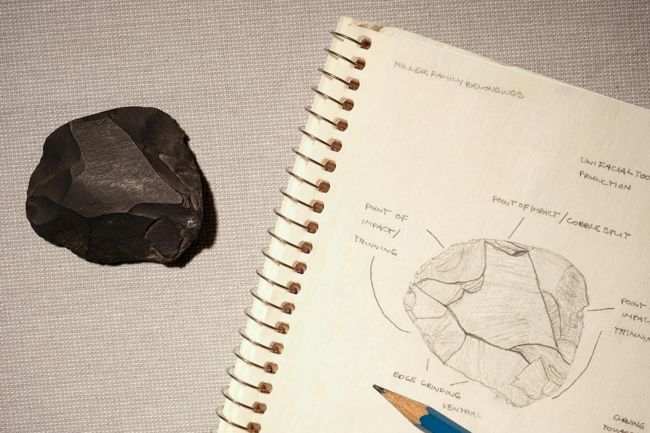 A rough black stone tool with sharp edges next to a spiral-bound notebook with a detailed pencil sketch of the stone, including annotations about impact points and edges.