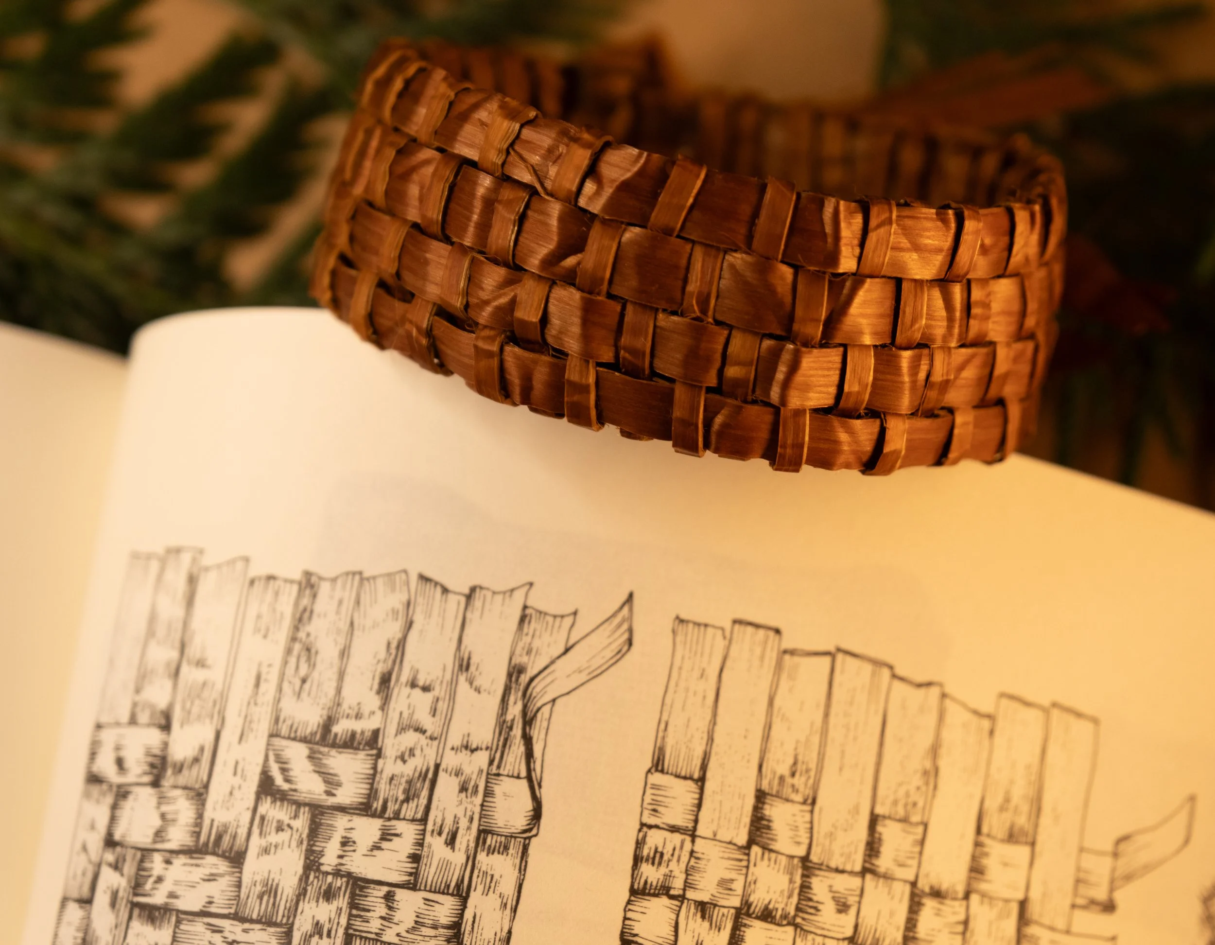 Close-up of a woven red cedar bracelet resting on an open reference book with black and white drawings of woven baskets.