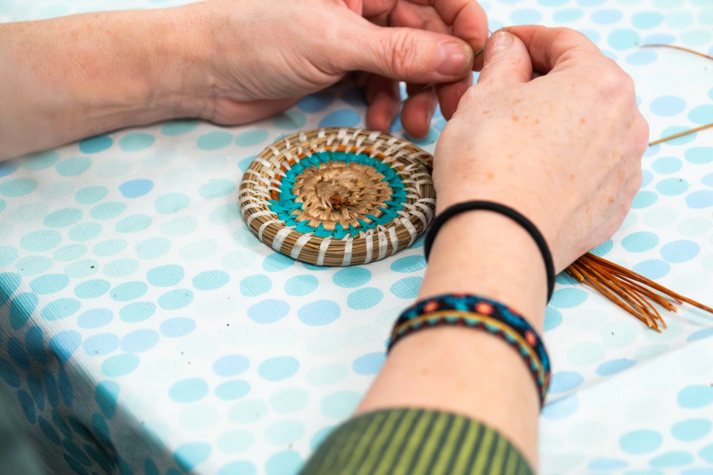 Person weaving a circular pine needle basket, sitting at a table with a blue polka-dot tablecloth.
