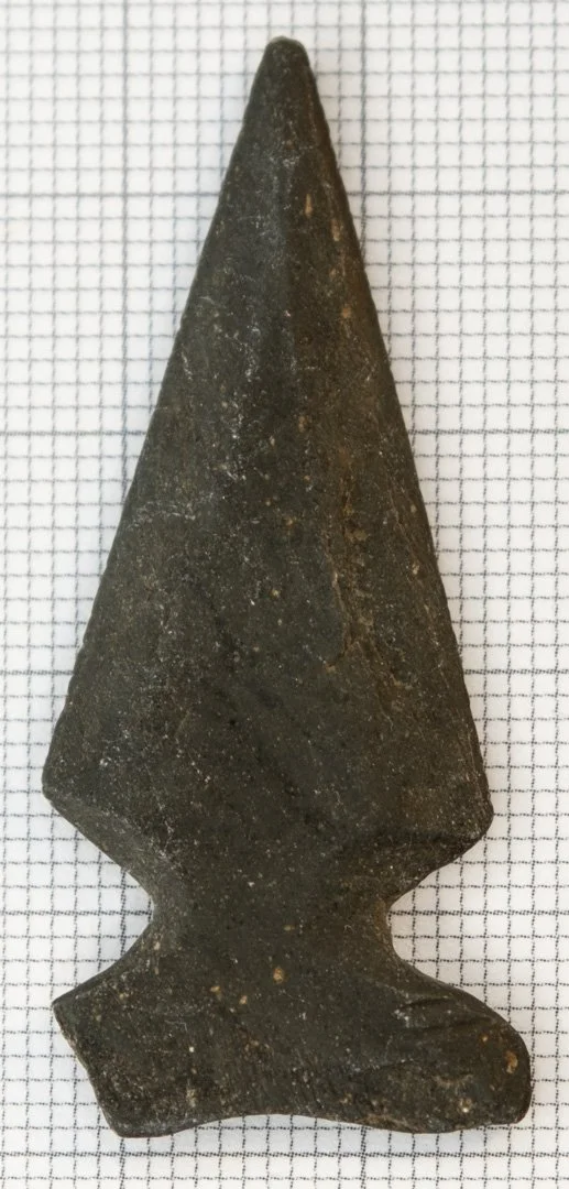 Dark grey slate projectile point on 1 mm graph paper for scale.